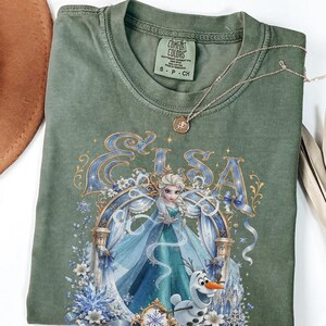 May include: An olive green t-shirt featuring a graphic print of the character Elsa from Frozen, with the name Elsa in large blue letters above her. The design includes a snowman, blue ribbons, white flowers, and snowflakes. The shirt has a Comfort Colors tag.