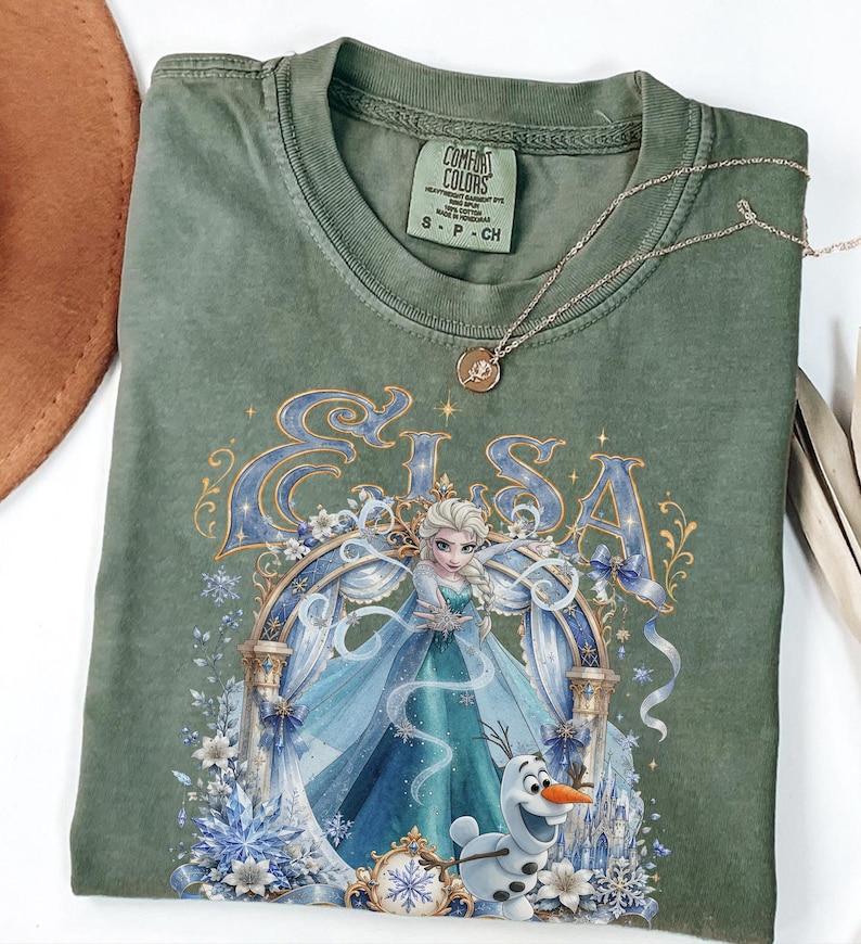 May include: An olive green t-shirt featuring a graphic print of the character Elsa from Frozen, with the name Elsa in large blue letters above her. The design includes a snowman, blue ribbons, white flowers, and snowflakes. The shirt has a Comfort Colors tag.