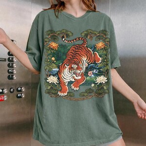 K&ouml;nnte beinhalten: Gr&uuml;nes kurz&auml;rmeliges T-Shirt mit einer detaillierten Tiger-Illustration. Der Tiger ist orange und schwarz, vor einem Hintergrund aus Blumen und Bl&auml;ttern. Das Design wird von &Auml;sten und Blumen umrahmt und schafft eine von der Natur inspirierte &Auml;sthetik.