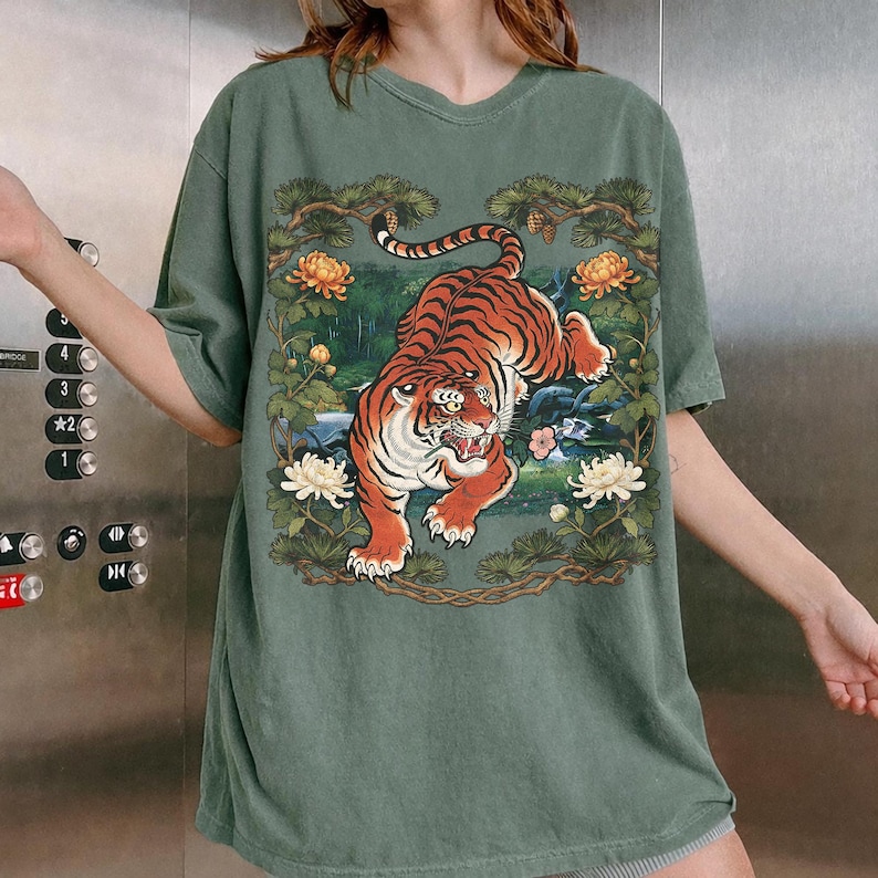 K&ouml;nnte beinhalten: Gr&uuml;nes kurz&auml;rmeliges T-Shirt mit einer detaillierten Tiger-Illustration. Der Tiger ist orange und schwarz, vor einem Hintergrund aus Blumen und Bl&auml;ttern. Das Design wird von &Auml;sten und Blumen umrahmt und schafft eine von der Natur inspirierte &Auml;sthetik.