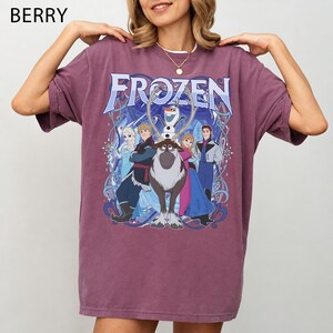 May include: A berry-colored short-sleeve t-shirt featuring a graphic print of the Frozen movie characters including Elsa, Anna, Kristoff, Olaf, and Sven. The text Frozen is printed in a large blue font above the characters with swirling blue accents.