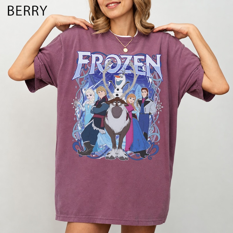 May include: A berry-colored short-sleeve t-shirt featuring a graphic print of the Frozen movie characters including Elsa, Anna, Kristoff, Olaf, and Sven. The text Frozen is printed in a large blue font above the characters with swirling blue accents.