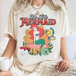 May include: A cream-colored short-sleeve t-shirt featuring a graphic print of The Little Mermaid. The design displays Ariel sitting in a large seashell with Flounder, Sebastian, and other sea creatures. The text reads The Little Mermaid.