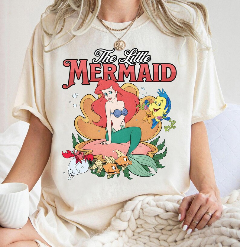 May include: A cream-colored short-sleeve t-shirt featuring a graphic print of The Little Mermaid. The design displays Ariel sitting in a large seashell with Flounder, Sebastian, and other sea creatures. The text reads The Little Mermaid.