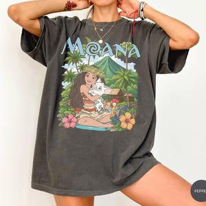 May include: A charcoal grey short-sleeve t-shirt featuring a graphic print of the character Moana holding a piglet, with a rooster, palm trees, and a mountain in the background. The text MOANA is printed in light blue above the illustration.