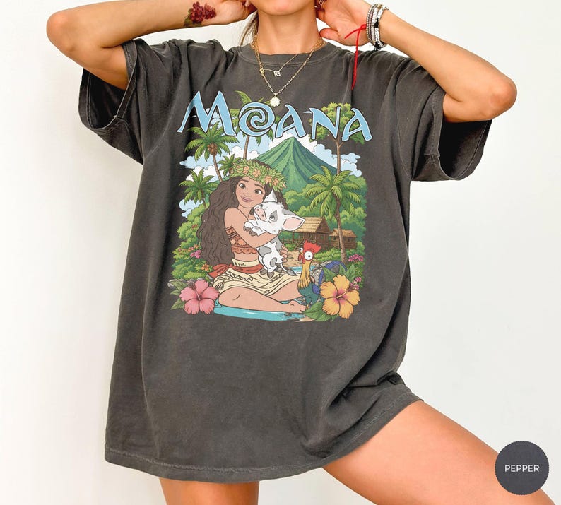 May include: A charcoal grey short-sleeve t-shirt featuring a graphic print of the character Moana holding a piglet, with a rooster, palm trees, and a mountain in the background. The text MOANA is printed in light blue above the illustration.