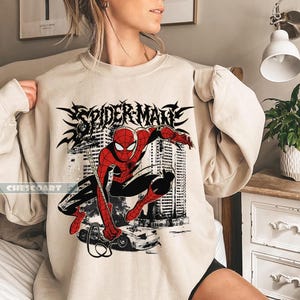 May include: Beige sweatshirt featuring a graphic of Spider-Man in a dynamic pose, with the words "SPIDER-MAN" in a stylized font. The design includes a cityscape and a car. The brand name "CHESCOART" is printed on the lower left.