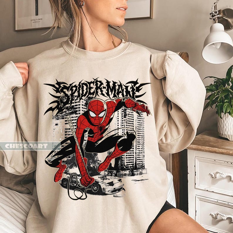 May include: Beige sweatshirt featuring a graphic of Spider-Man in a dynamic pose, with the words "SPIDER-MAN" in a stylized font. The design includes a cityscape and a car. The brand name "CHESCOART" is printed on the lower left.