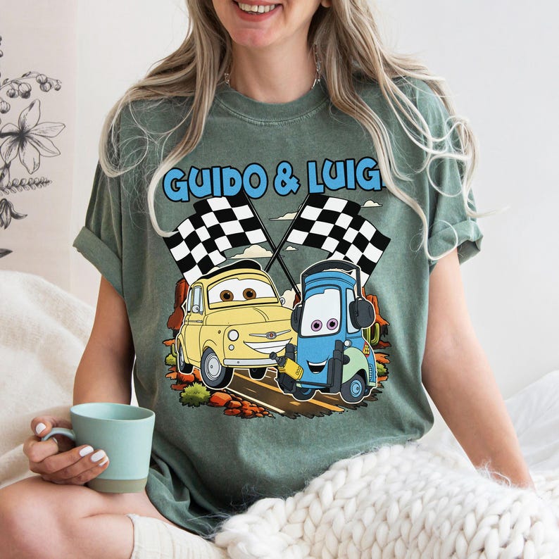 May include: Green t-shirt featuring the text "GUIDO & LUIG" above a cartoon image of two race cars with checkered flags. One car is yellow, the other is blue. The shirt is a comfortable, casual style.