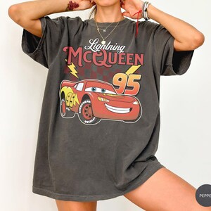 May include: A charcoal gray short-sleeve t-shirt featuring a graphic of the red race car Lightning McQueen with the text Lightning MCQUEEN and the number 95. The design includes a yellow lightning bolt and a dark red checkered pattern background.