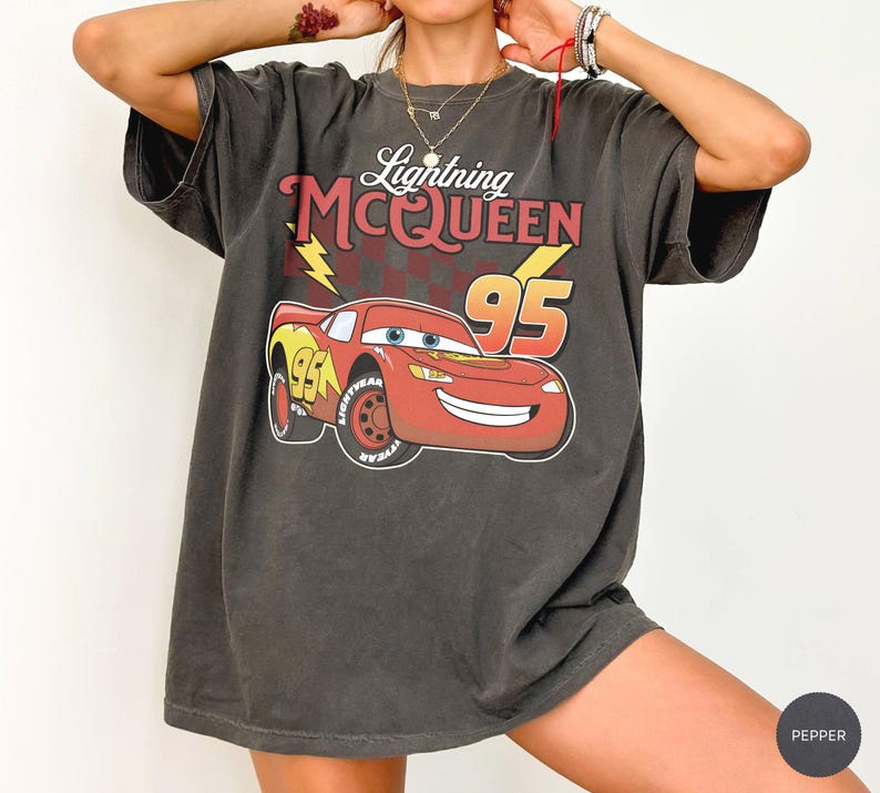 May include: A charcoal gray short-sleeve t-shirt featuring a graphic of the red race car Lightning McQueen with the text Lightning MCQUEEN and the number 95. The design includes a yellow lightning bolt and a dark red checkered pattern background.