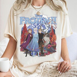 May include: A cream-colored short-sleeve t-shirt featuring a graphic print of the Disney Frozen II movie characters Anna, Elsa, Kristoff, Sven, and Olaf. The design includes a mountain landscape with autumn trees and the Frozen II logo text.