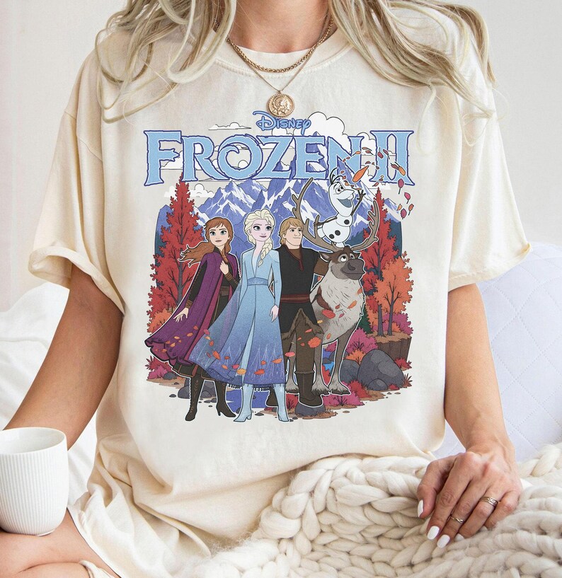 May include: A cream-colored short-sleeve t-shirt featuring a graphic print of the Disney Frozen II movie characters Anna, Elsa, Kristoff, Sven, and Olaf. The design includes a mountain landscape with autumn trees and the Frozen II logo text.
