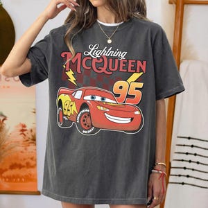 May include: A charcoal gray short-sleeve t-shirt featuring a graphic of the red race car Lightning McQueen with the number 95, a yellow lightning bolt, and a checkered flag pattern. The text reads Lightning McQUEEN above the car illustration.