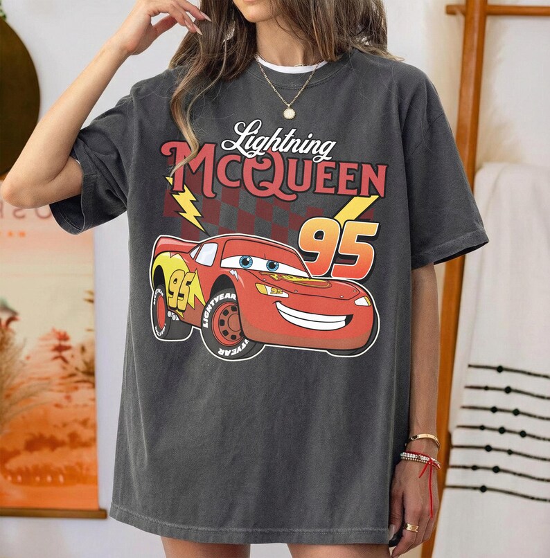 May include: A charcoal gray short-sleeve t-shirt featuring a graphic of the red race car Lightning McQueen with the number 95, a yellow lightning bolt, and a checkered flag pattern. The text reads Lightning McQUEEN above the car illustration.