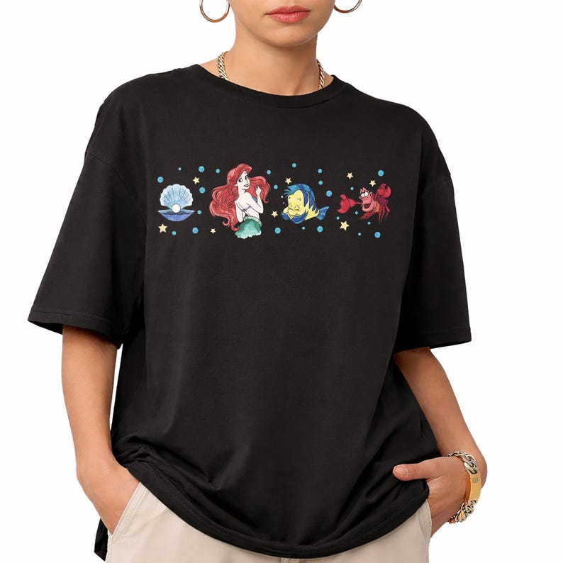 K&ouml;nnte beinhalten: Schwarzes T-Shirt mit einer farbenfrohen Grafik von Figuren aus einem beliebten Zeichentrickfilm. Das Design umfasst eine Muschel, eine Meerjungfrau mit roten Haaren, einen gelben Fisch und eine rote Krabbe mit blauen Punkten und gelben Sternen.