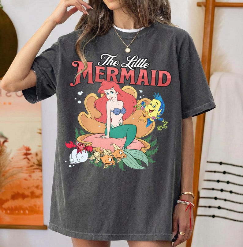 May include: A charcoal gray t-shirt featuring a graphic print of The Little Mermaid with Ariel sitting in a large seashell, accompanied by Flounder and Sebastian. The text The Little Mermaid is printed in red above the character illustration.