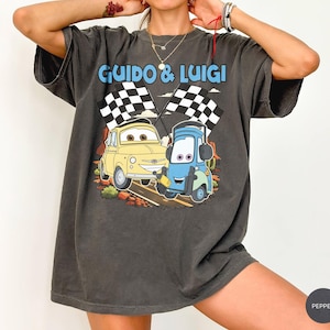 May include: Dark gray t-shirt featuring the text "Guido & Luigi" above a cartoon image of two race cars with checkered flags. The yellow car is smiling, and the blue car is holding a wrench. The shirt has a vintage, worn look.