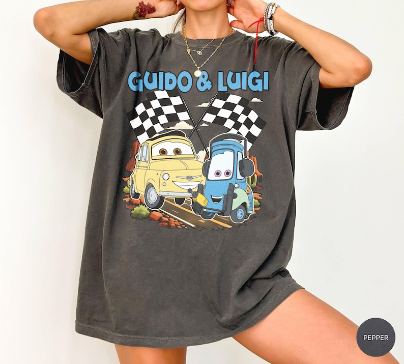 May include: Dark gray t-shirt featuring the text "Guido & Luigi" above a cartoon image of two race cars with checkered flags. The yellow car is smiling, and the blue car is holding a wrench. The shirt has a vintage, worn look.