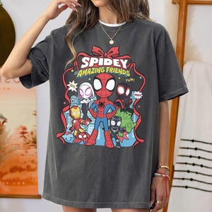 May include: A charcoal grey short-sleeve t-shirt featuring a graphic print of Spidey and His Amazing Friends. The design includes cartoon versions of Spider-Man, Iron Man, Hulk, and other characters framed by a red ribbon with the text Spidey and His Amazing Friends.