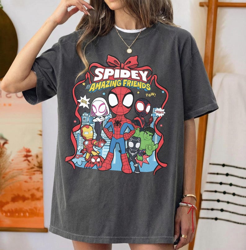 May include: A charcoal grey short-sleeve t-shirt featuring a graphic print of Spidey and His Amazing Friends. The design includes cartoon versions of Spider-Man, Iron Man, Hulk, and other characters framed by a red ribbon with the text Spidey and His Amazing Friends.