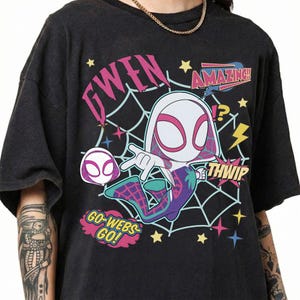 May include: A black t-shirt featuring a graphic of a cartoon character in a white, pink, and purple suit with a spiderweb background. The text reads GWEN, AMAZING, THWIP, and GO-WEBS GO! along with yellow stars and lightning bolt accents.
