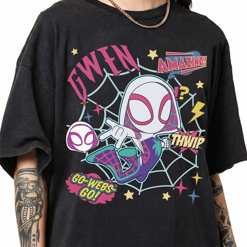 May include: A black t-shirt featuring a graphic of a cartoon character in a white, pink, and purple suit with a spiderweb background. The text reads GWEN, AMAZING, THWIP, and GO-WEBS GO! along with yellow stars and lightning bolt accents.