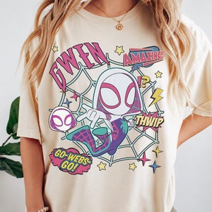 May include: A cream-colored t-shirt featuring a graphic of a cartoon character in a white and pink hooded suit with a spiderweb background. The text reads GWEN, AMAZING, THWIP, and GO-WEBS-GO, accompanied by yellow stars, lightning bolts, and sparkles.