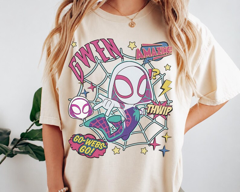 May include: A cream-colored t-shirt featuring a graphic of a cartoon character in a white and pink hooded suit with a spiderweb background. The text reads GWEN, AMAZING, THWIP, and GO-WEBS-GO, accompanied by yellow stars, lightning bolts, and sparkles.