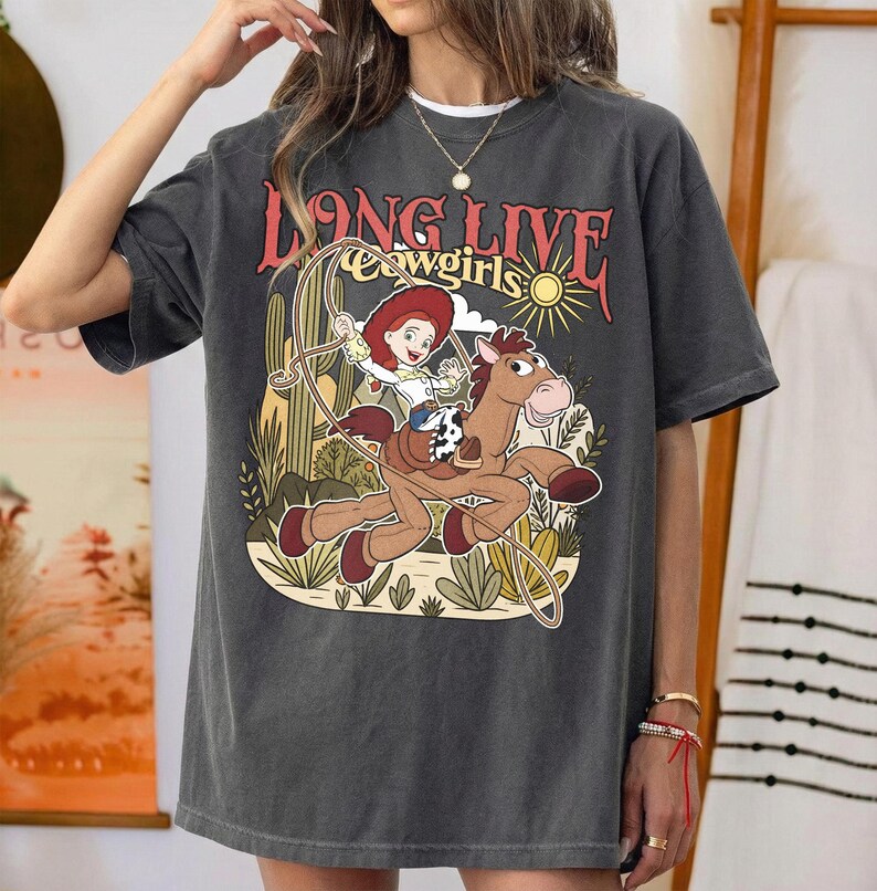 Puede incluir: Una camiseta gris carb&oacute;n de manga corta con un estampado gr&aacute;fico de una vaquera montando un caballo con un lazo. El texto dice LONG LIVE cowgirls. El dise&ntilde;o incluye un paisaje des&eacute;rtico con cactus, un sol y tonos tierra apagados.