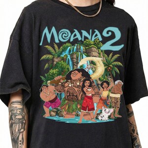 May include: A black short-sleeve t-shirt featuring a graphic print of characters from Moana 2. The design includes the text Moana 2 in light blue, a waterfall, palm trees, and a group of animated characters including a large figure holding a hook.