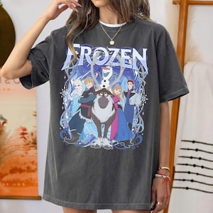May include: A charcoal gray short-sleeve t-shirt featuring a large graphic print of the characters Elsa, Anna, Kristoff, Olaf, and Sven from the movie Frozen. The text Frozen is printed in a large blue font above the character illustration.