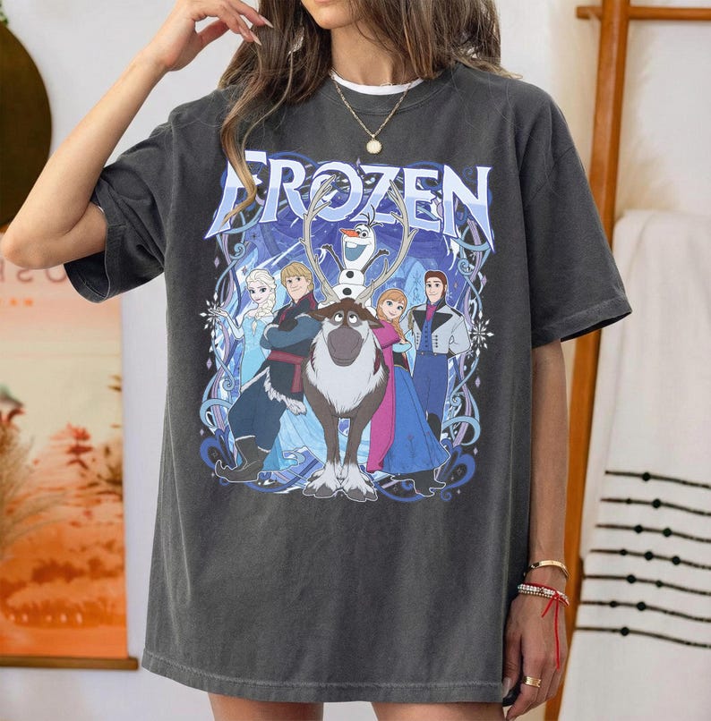 May include: A charcoal gray short-sleeve t-shirt featuring a large graphic print of the characters Elsa, Anna, Kristoff, Olaf, and Sven from the movie Frozen. The text Frozen is printed in a large blue font above the character illustration.