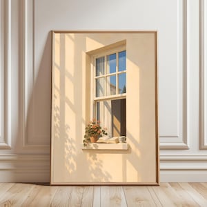 May include: A framed wall art print depicting a beige wall with a window. On the window sill sits a potted plant with orange flowers, an open book, and a small mug. Sunlight casts long shadows across the wall, creating a warm atmosphere.