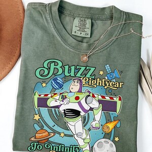 May include: An olive green t-shirt featuring a graphic of Buzz Lightyear with the text Buzz Lightyear To Infinity. The design includes space elements like planets, stars, a satellite, and a comet. The shirt has a Comfort Colors brand label.