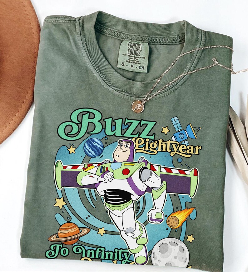 May include: An olive green t-shirt featuring a graphic of Buzz Lightyear with the text Buzz Lightyear To Infinity. The design includes space elements like planets, stars, a satellite, and a comet. The shirt has a Comfort Colors brand label.