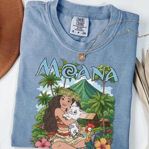 May include: A blue short-sleeved t-shirt featuring a graphic print of the character Moana holding a piglet, with a rooster, tropical palm trees, a mountain, and the text MOANA. The shirt has a crew neckline and a Comfort Colors brand label inside.