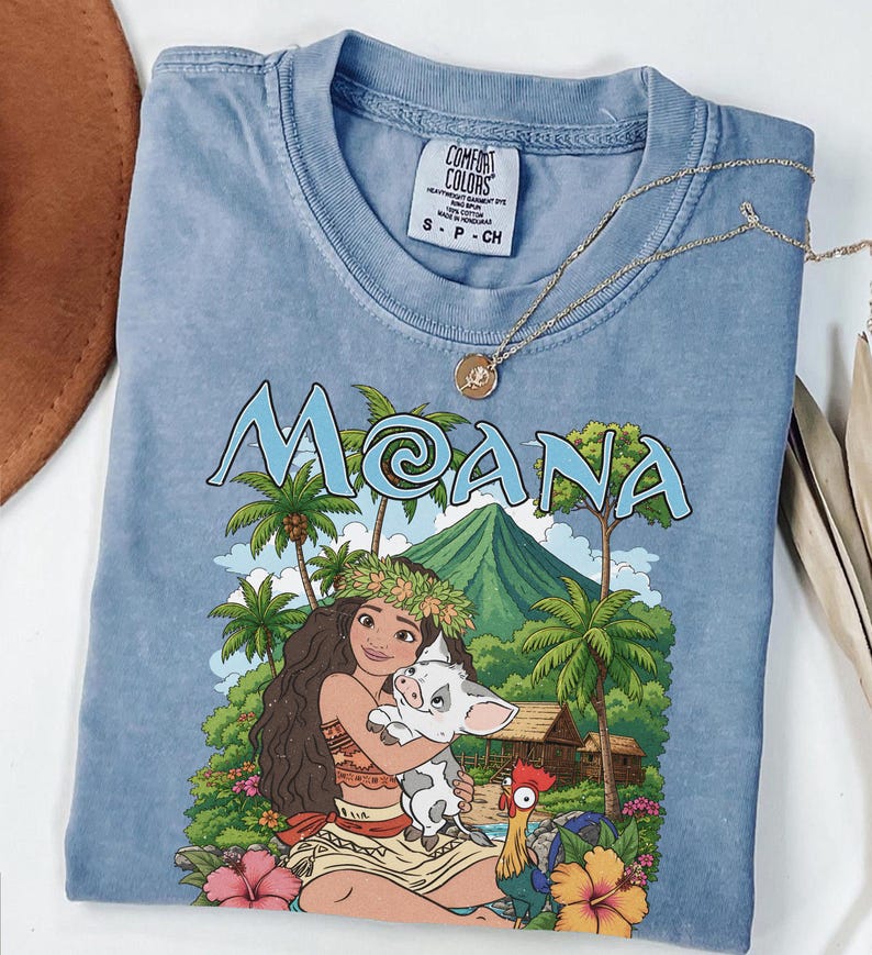 May include: A blue short-sleeved t-shirt featuring a graphic print of the character Moana holding a piglet, with a rooster, tropical palm trees, a mountain, and the text MOANA. The shirt has a crew neckline and a Comfort Colors brand label inside.