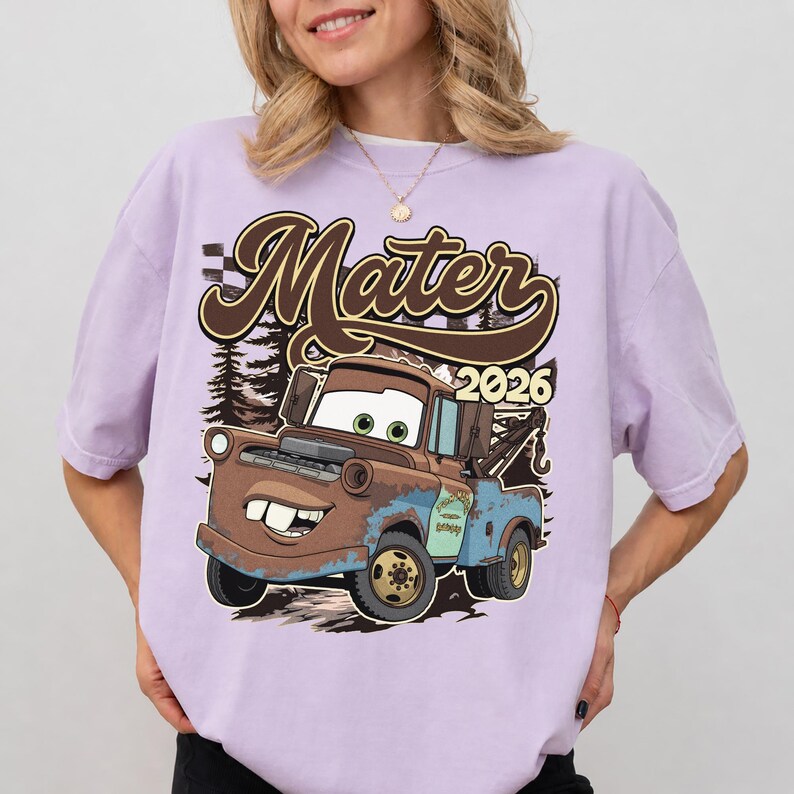 May include: A light purple t-shirt featuring a cartoon image of Mater from the Cars movie. The graphic includes the name "Mater" in brown script, the year "2026", and a backdrop of trees. The tow truck Mater is brown and blue.