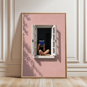May include: A framed wall art print featuring a pink wall with an open white window. A person sits in the window frame reading a blue book. The scene includes shadows of tree leaves cast onto the pink wall, creating a contrast between light and dark.