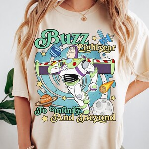 May include: A cream-colored t-shirt featuring a graphic of Buzz Lightyear in a space setting with planets, stars, and a satellite. The text reads Buzz Lightyear To Infinity And Beyond in a retro font style with blue, green, and yellow colors.