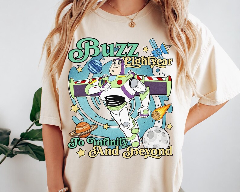 May include: A cream-colored t-shirt featuring a graphic of Buzz Lightyear in a space setting with planets, stars, and a satellite. The text reads Buzz Lightyear To Infinity And Beyond in a retro font style with blue, green, and yellow colors.