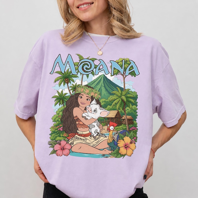 May include: A light purple t-shirt featuring a graphic print of a long-haired character holding a small pig, with a rooster nearby. The scene includes palm trees, a mountain, and a hut. The text MOANA is printed in blue at the top of the graphic.