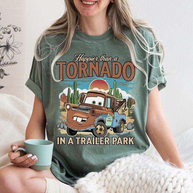 May include: Green t-shirt featuring a cartoon tow truck in a desert landscape. The text on the shirt says "Happier than a Tornado in a Trailer Park". The shirt has rolled sleeves and a casual fit.