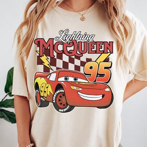 May include: A cream-colored t-shirt featuring a graphic of the red race car Lightning McQueen with the number 95, a checkered flag background, and yellow lightning bolts. The text Lightning MCQUEEN is printed in red above the car graphic.