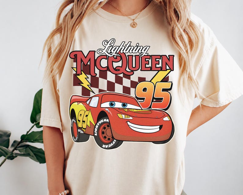 May include: A cream-colored t-shirt featuring a graphic of the red race car Lightning McQueen with the number 95, a checkered flag background, and yellow lightning bolts. The text Lightning MCQUEEN is printed in red above the car graphic.