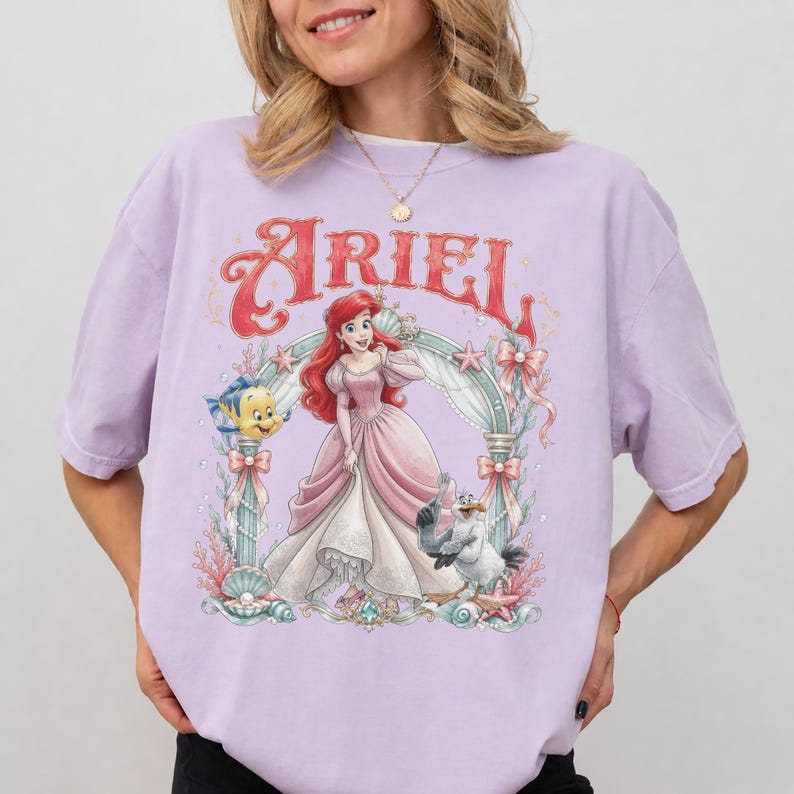 May include: A light purple short-sleeve t-shirt featuring a graphic print of Ariel in a pink gown, surrounded by an ornate archway with Flounder and Scuttle. The text Ariel is printed in a large red decorative font above the character illustration.