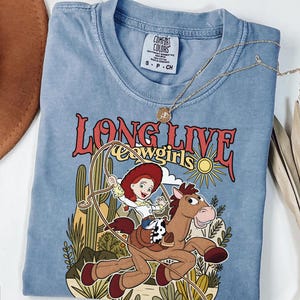 Puede incluir: Una camiseta azul de manga corta con un gr&aacute;fico de una vaquera montando un caballo marr&oacute;n con un lazo. El texto dice LONG LIVE cowgirls. El dise&ntilde;o incluye cactus, un sol y nubes. Etiqueta de Comfort Colors en el cuello.
