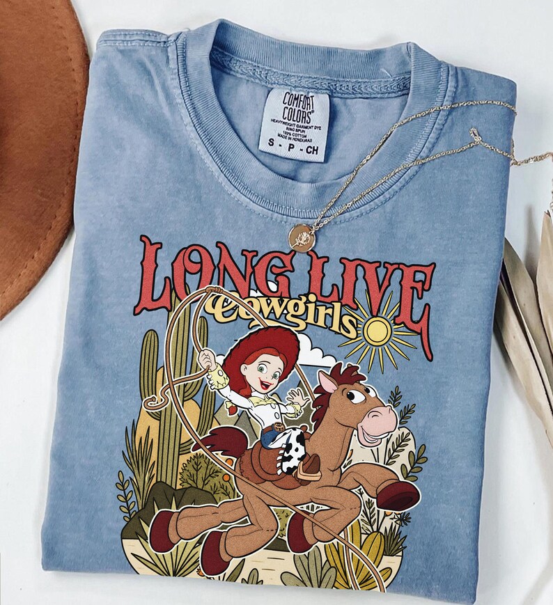 Puede incluir: Una camiseta azul de manga corta con un gr&aacute;fico de una vaquera montando un caballo marr&oacute;n con un lazo. El texto dice LONG LIVE cowgirls. El dise&ntilde;o incluye cactus, un sol y nubes. Etiqueta de Comfort Colors en el cuello.