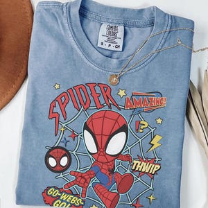 May include: A blue short-sleeved t-shirt featuring a graphic of a cartoon spider character in a red and blue suit. The design includes a spider web background, yellow stars, and text that reads SPIDER, AMAZING, THWIP, and GO-WEBS-GO.
